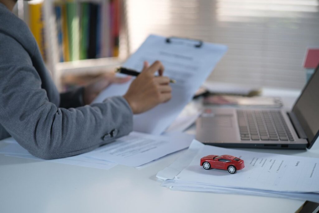 A person writes on a clipboard in front of a laptop at a desk next to a red model car.