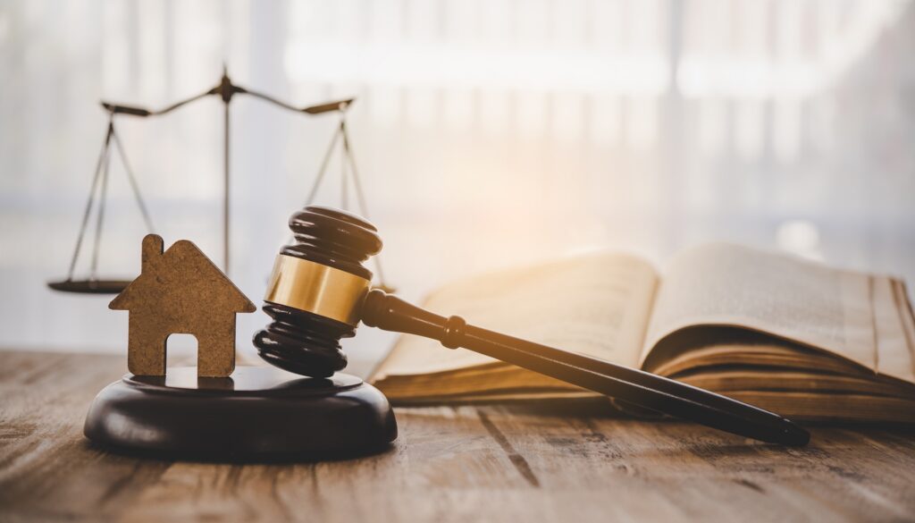 A gavel rests on its stand next to a small house and in front of a book and scales of justice.
