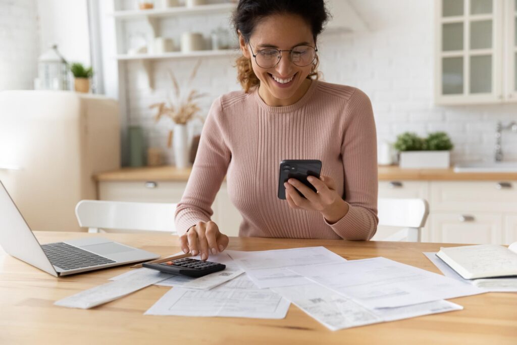 A woman smiles at her cell phone at a desk in front of paperwork and a laptop.