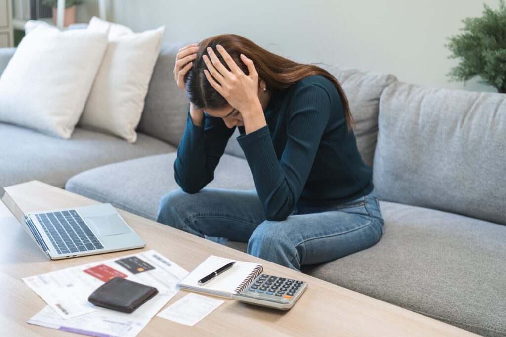 woman sitting on couch with her hands on her head in front of a table with a calculator and papers