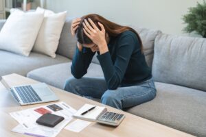 woman sitting on couch with her hands on her head in front of a table with a calculator and papers